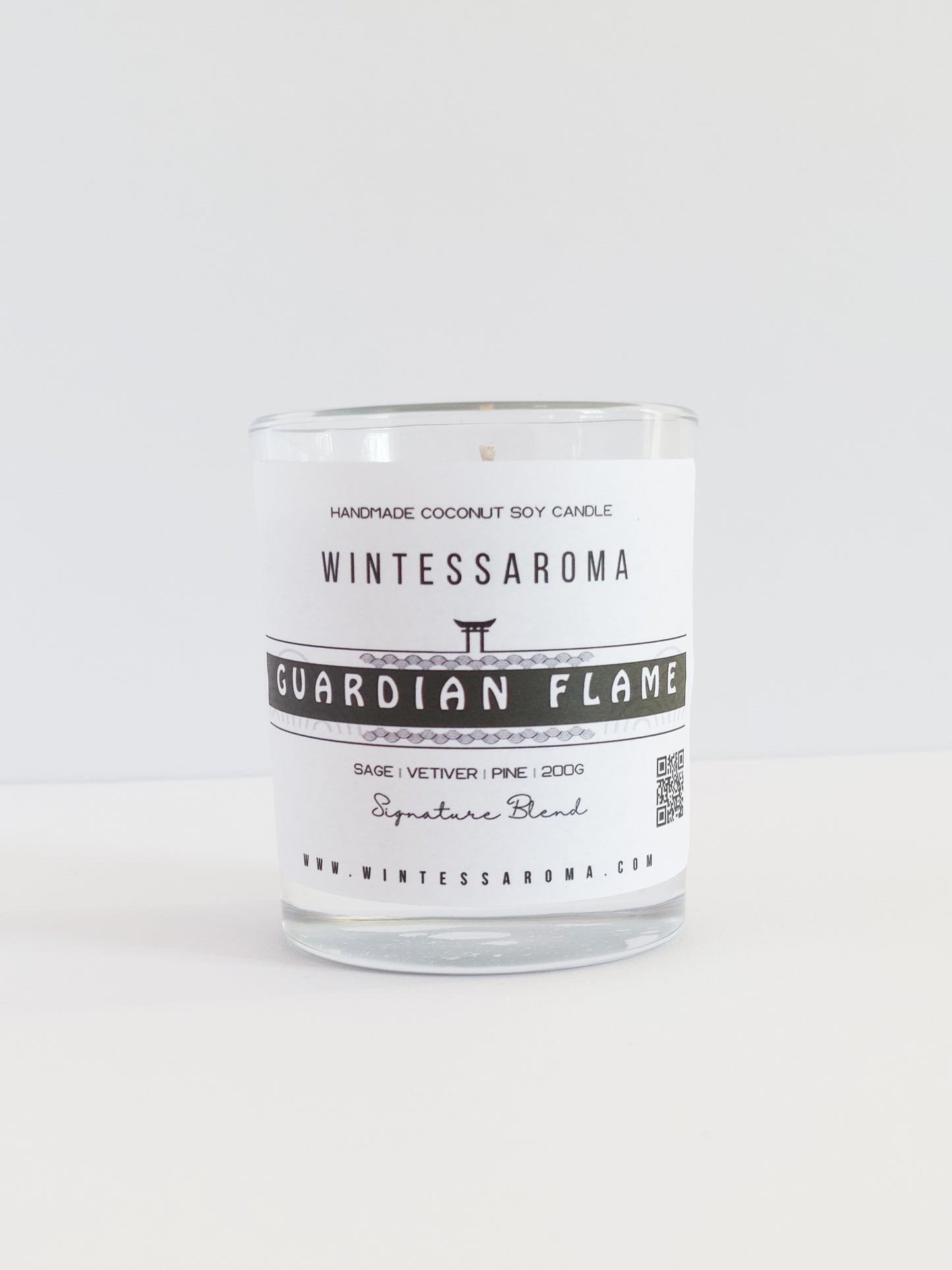 A clear glass candle with a label that reads 'WINTESSARAOMA GUARDIAN FLAME' The candle is placed on a white surface against a white background.