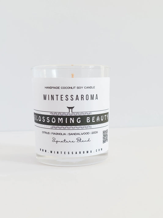 A clear glass candle with a label that reads 'Wintessaroma Blossoming Beauty Korean Tale'. The candle is placed on a white surface against a white background.