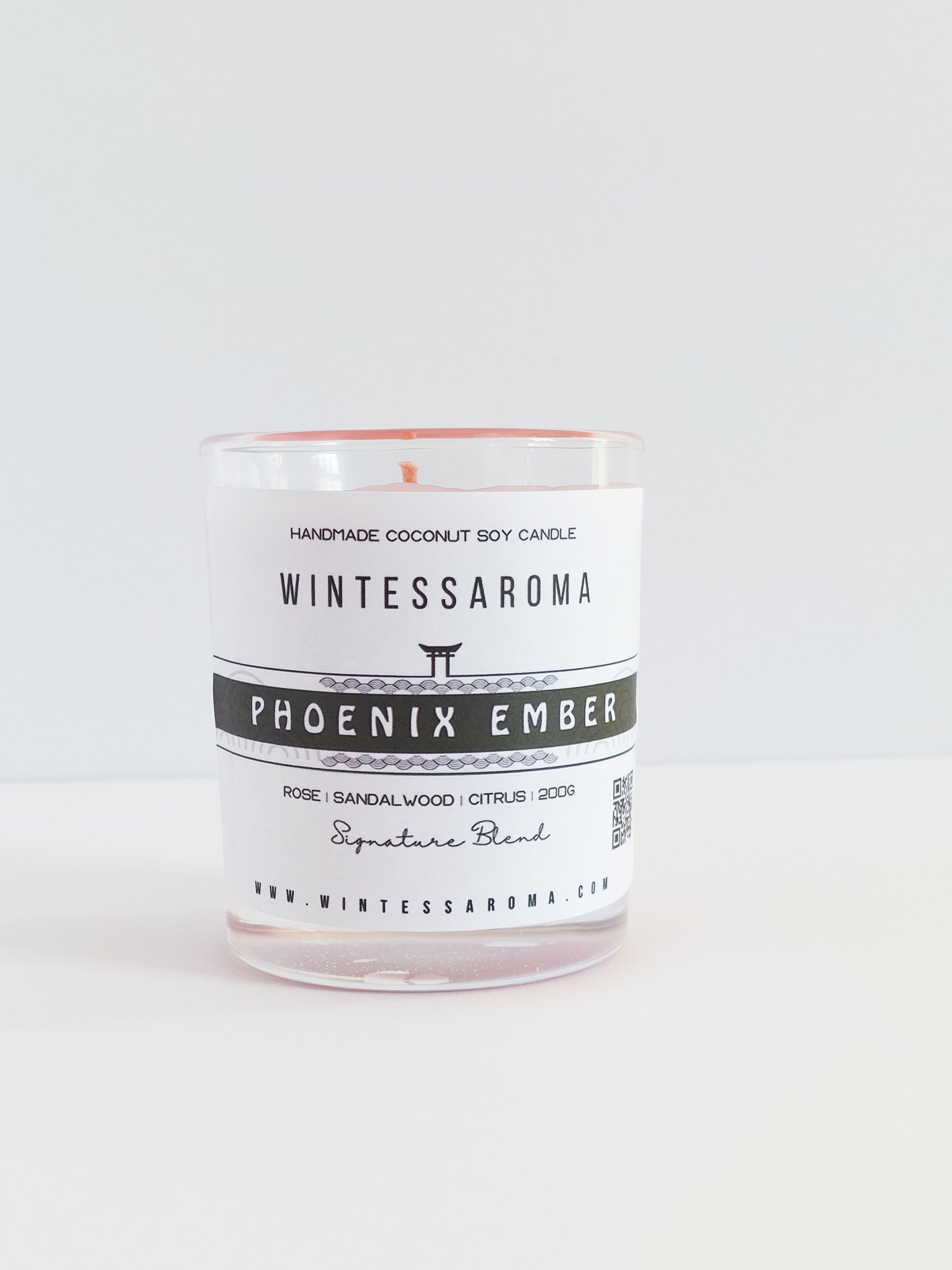 A clear glass candle with a white label that reads 'WINTESSAROMA PHOENIX EMBER' and a small amount of wax visible at the top.