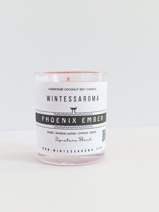 A clear glass candle with a white label that reads 'WINTESSAROMA PHOENIX EMBER' and a small amount of wax visible at the top.