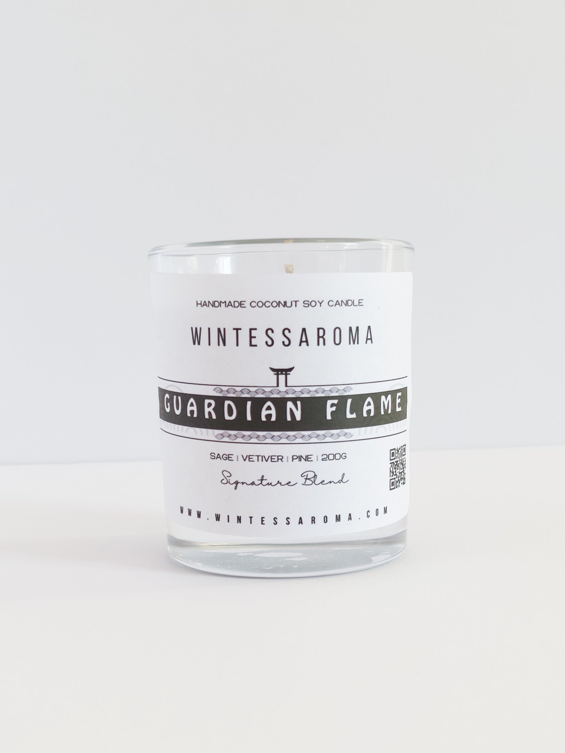 A clear glass candle with a label that reads 'WINTESSARAOMA GUARDIAN FLAME' The candle is placed on a white surface against a white background.