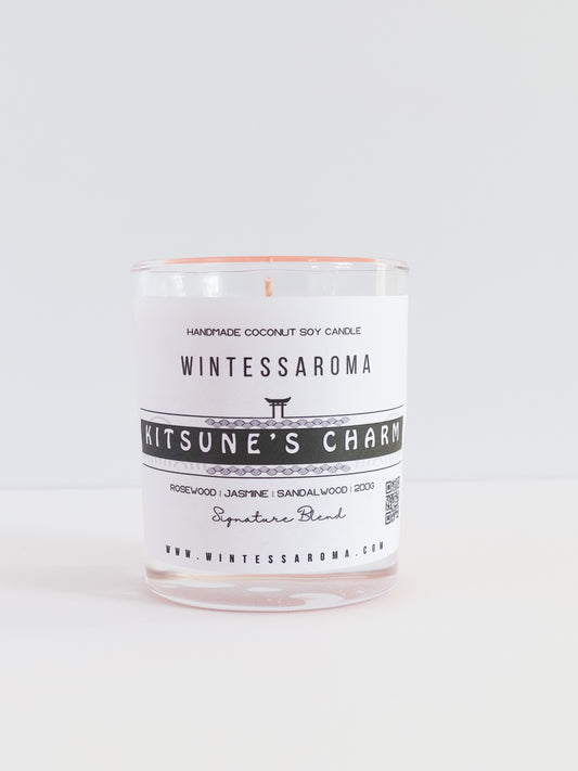 A clear glass candle with a label that reads 'Wintessaroma Kitsune's Charm' and is infused with floral and wood scents.