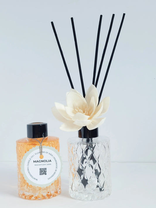 A reed diffuser with a clear glass base and a bottle labeled 'MAGNOLIA' in front of it, alongside a floral arrangement with white flowers and green leaves.