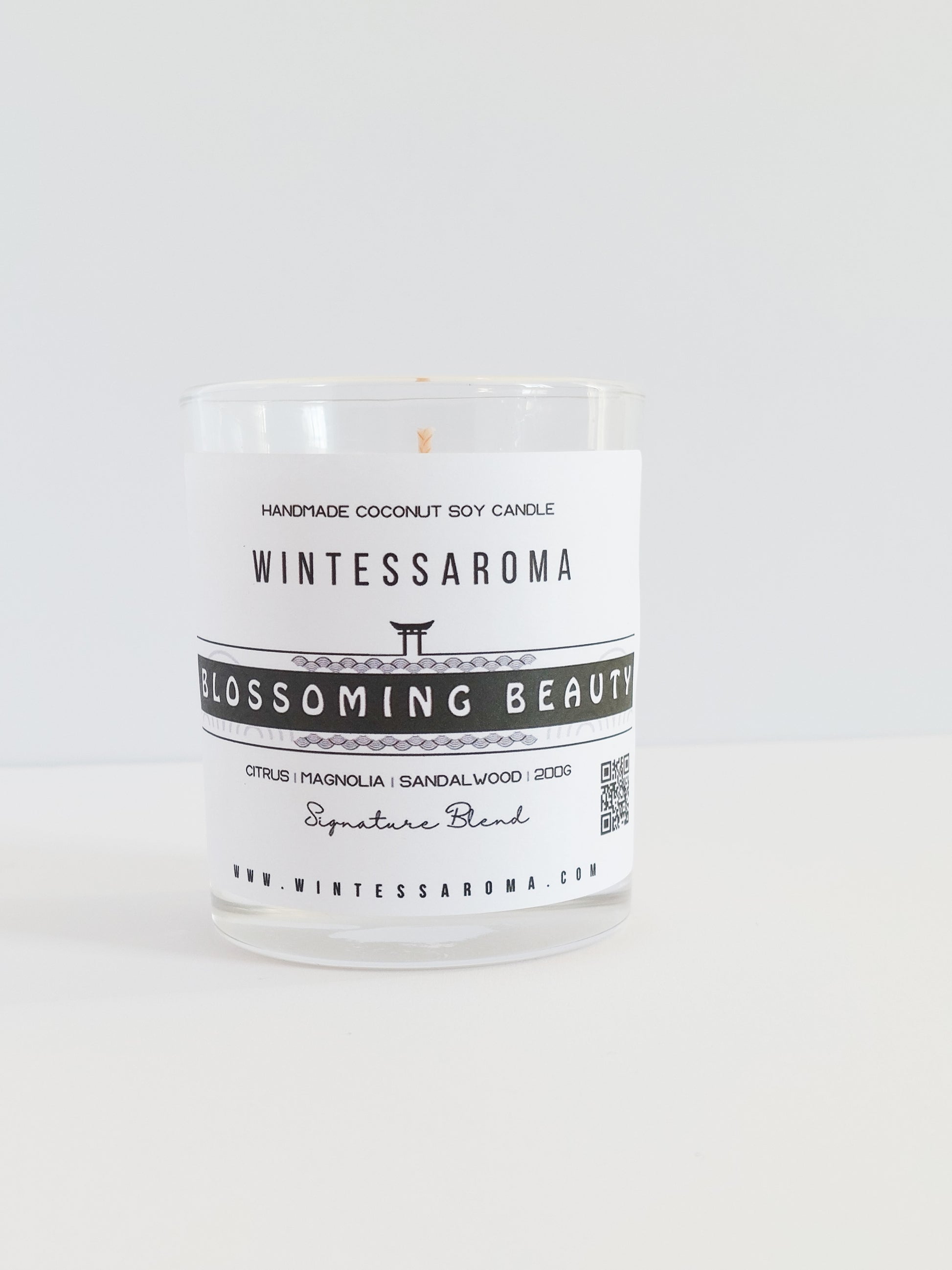 A clear glass candle with a label that reads 'Wintessaroma Blossoming Beauty Korean Tale'. The candle is placed on a white surface against a white background.