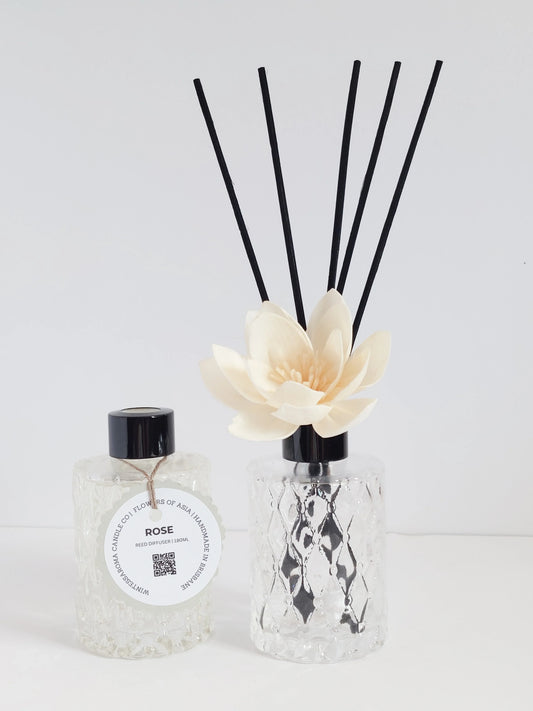 A beige reed diffuser with a clear glass top and a black spray bottle labeled 'ROSE', placed on a white surface.